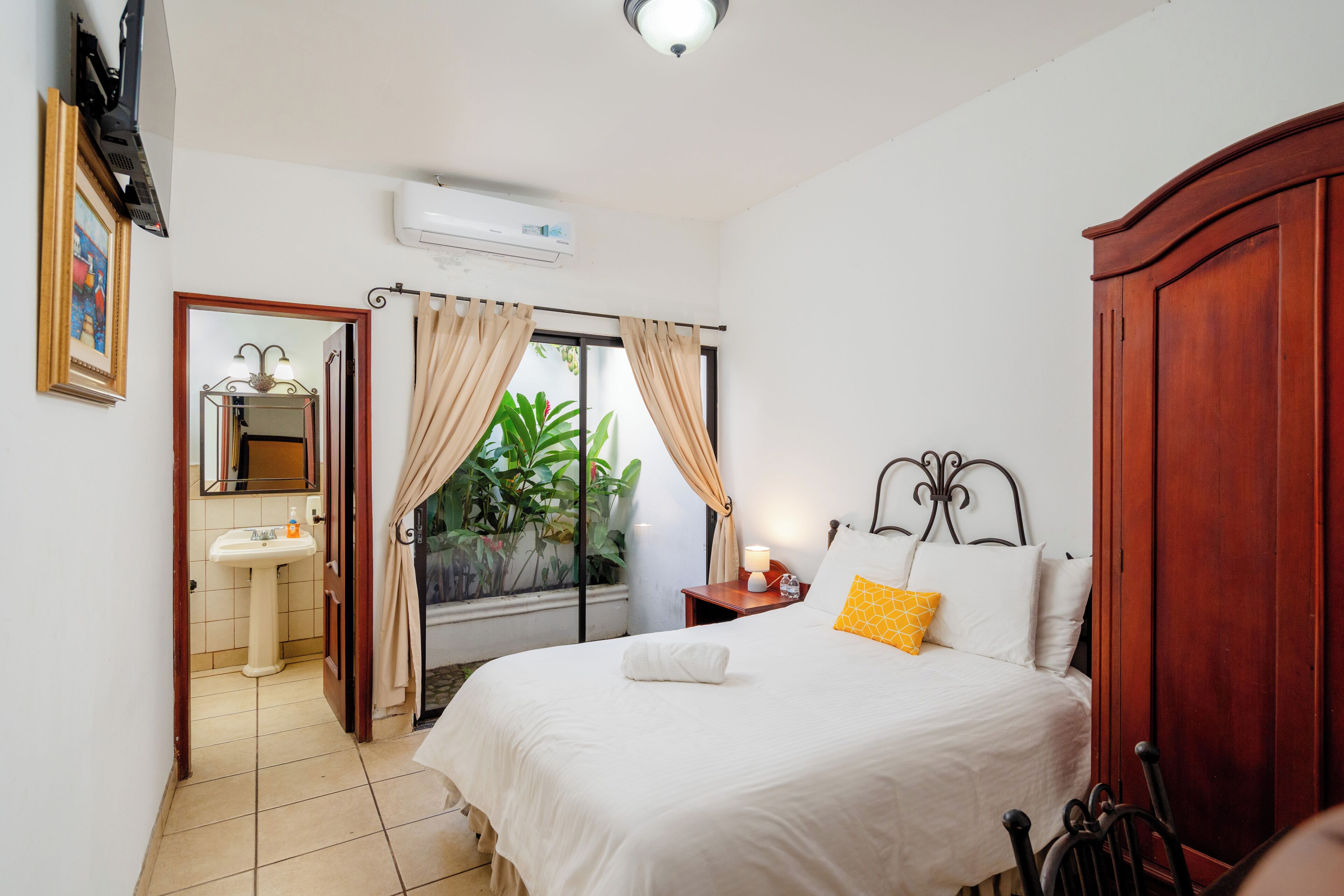 A bedroom with a bed, a nightstand, a bedside table, a window with curtains, a mirror, a sink, and a door leading to another room.