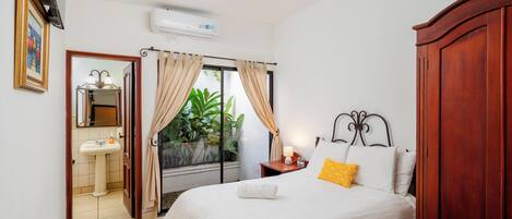 Un dormitorio con cama, mesita de noche, tocador, ventana con cortinas, espejo, lavamanos y puerta que da a otra habitación.