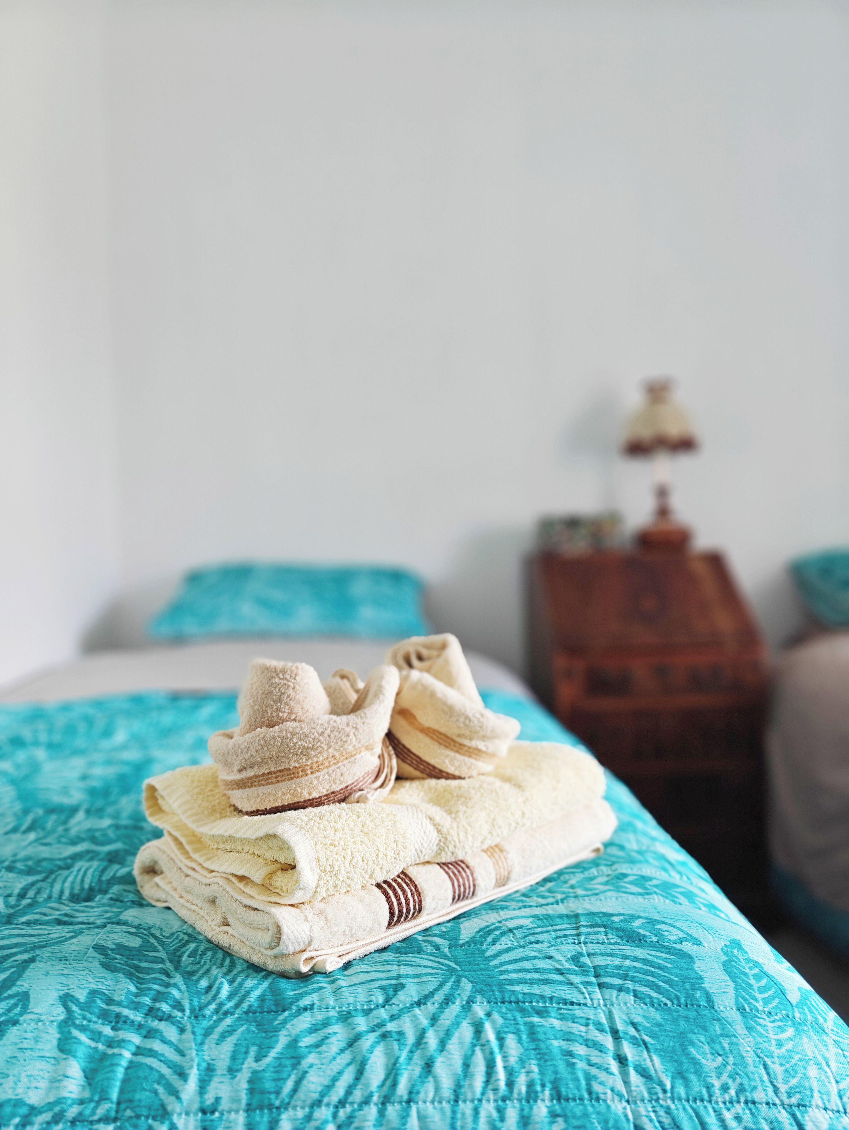 Una cama bien tendida con una colcha azul estampada, una toalla doblada y un par de zapatillas encima.