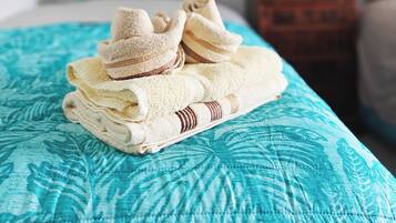 Una cama bien tendida con una colcha azul estampada, una toalla doblada y un par de zapatillas encima.
