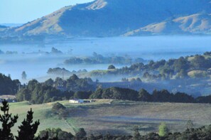 A rural landscape with rolling hills, a misty lake, and a small white building in the foreground. - Top Hill House (Maungatapere)