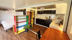 Une pièce de taille modeste comprenant un lit, une table à manger en bois, un coin cuisine avec des meubles noirs, un évier et une plante en pot.