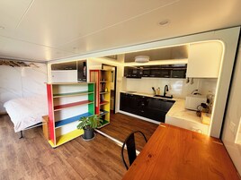 Una habitación pequeña con cama, una mesa de comedor de madera, un área de cocina con armarios negros, un lavamanos y una planta en maceta.