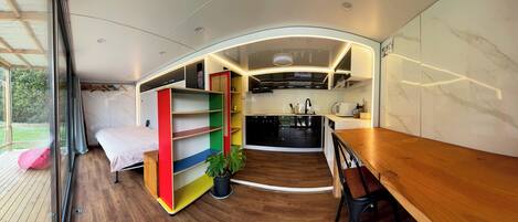 A compact, modern interior with a bed, a colorful bookshelf, a kitchen area, and a dining table.