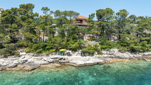 Una propiedad costera con costa rocosa, agua turquesa cristalina y un edificio rodeado de densos pinos.