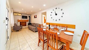 Comedor con mesa de madera dispuesta para cuatro, un reloj de pared grande y una cocina al fondo.