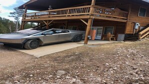 A two-story wooden cabin with a balcony, a car parked in front, and a gravel driveway. - {Aspen Chalet} Hot Tub, Tesla Charger, Near Deadwood! (Lead)