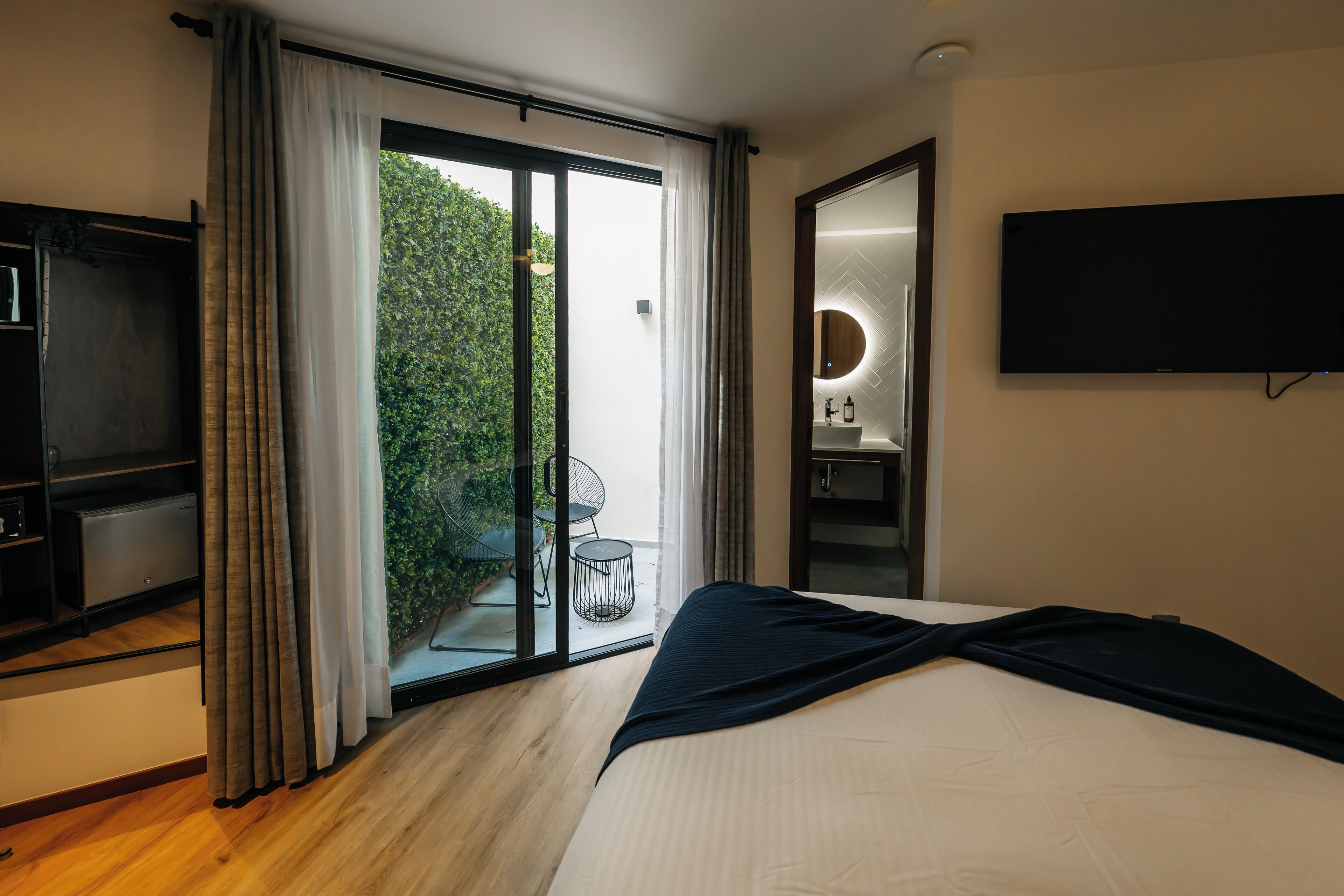 Een moderne hotelkamer met een groot bed, een flatscreen televisie, een glazen schuifdeur naar een balkon, en een klein rond tafeltje met stoelen.