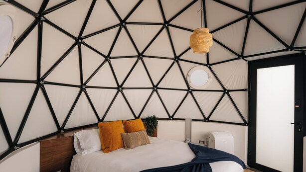 Una habitación tipo tienda de campaña, moderna, con diseño geométrico, que incluye una cama, una mesita auxiliar pequeña y un aplique colgante.