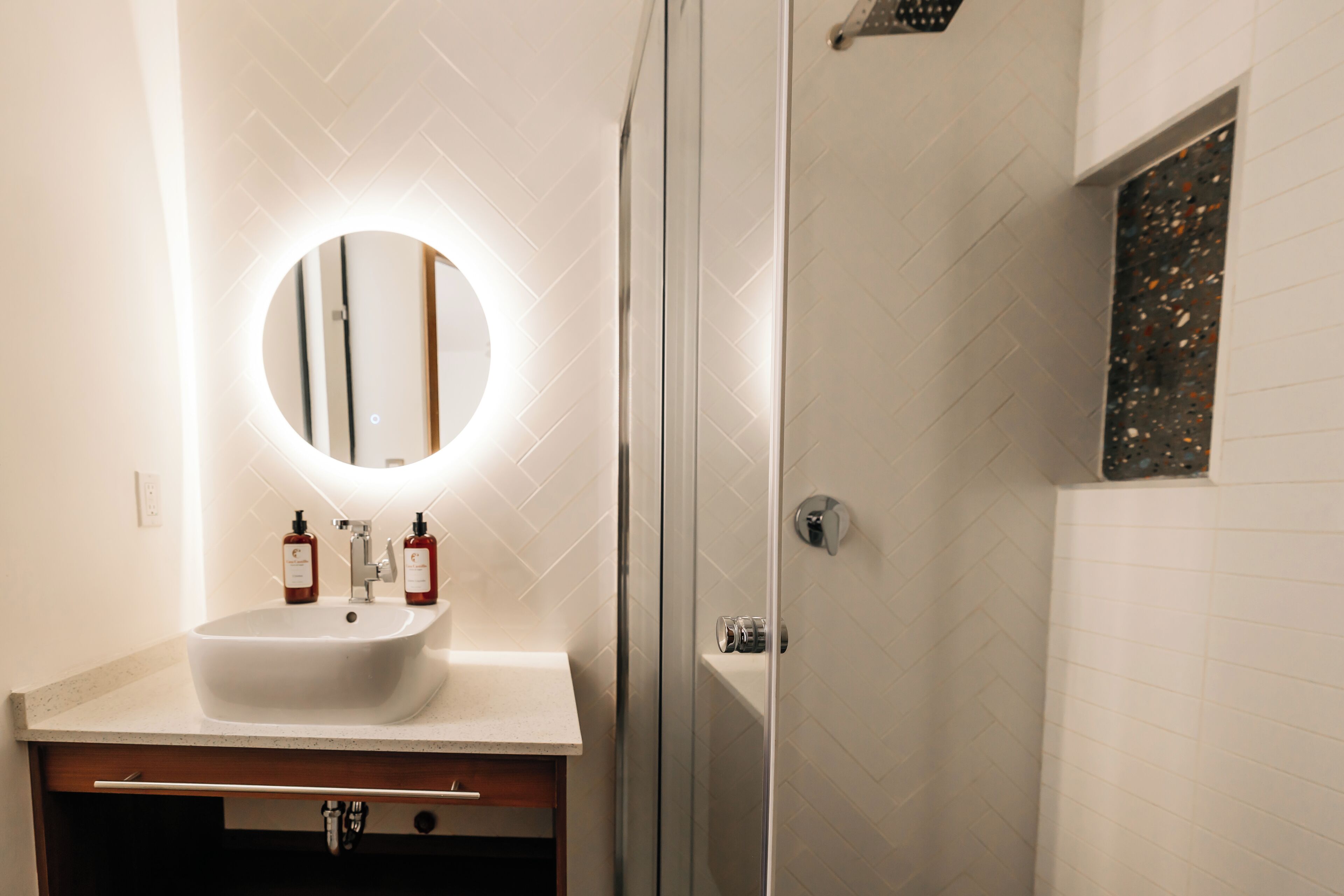 Une salle de bain avec un lavabo blanc, deux bouteilles de savon et un miroir à cadre rond.