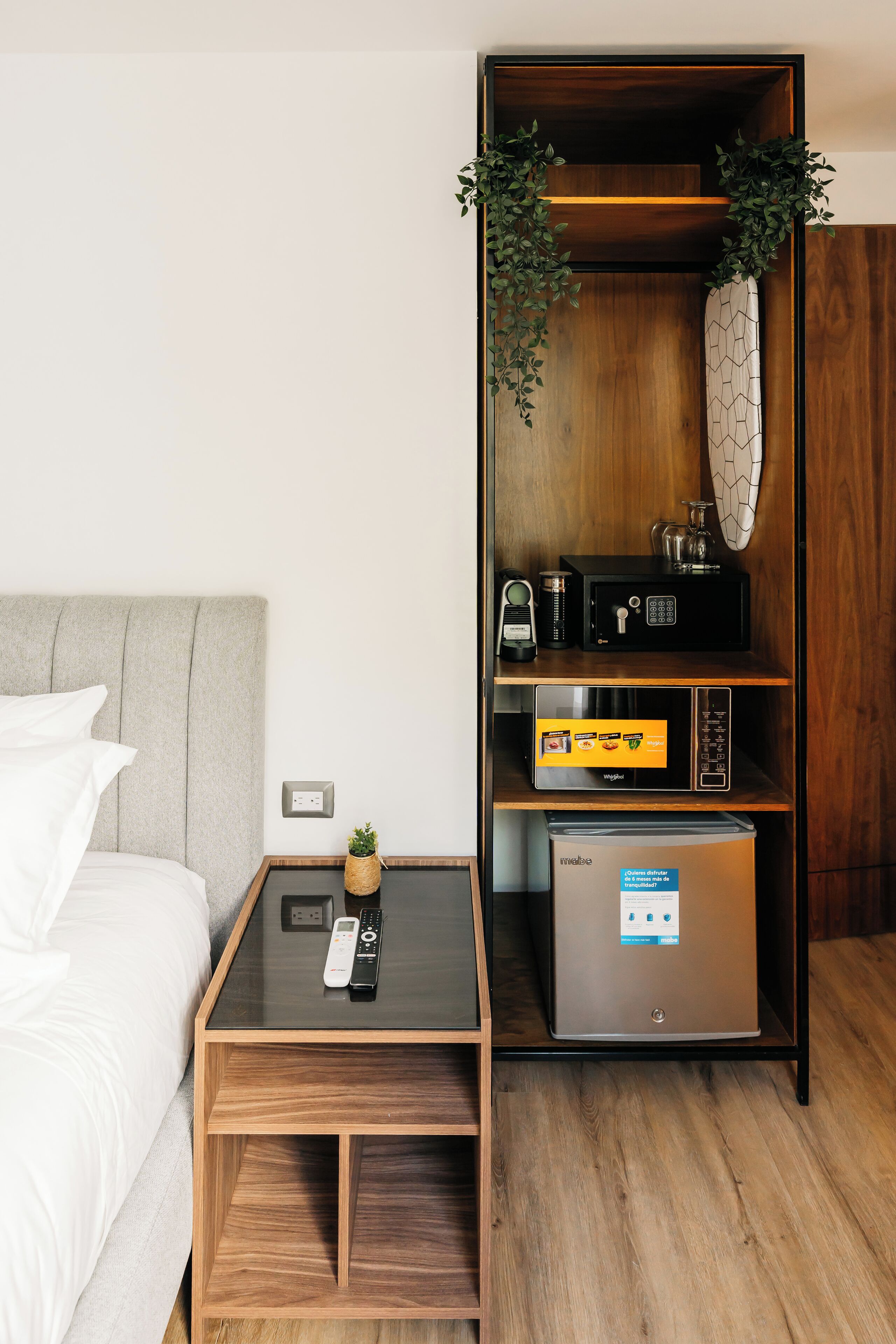 A hotel room with a bed, a bedside table with a remote control, a minibar, and a built-in refrigerator.