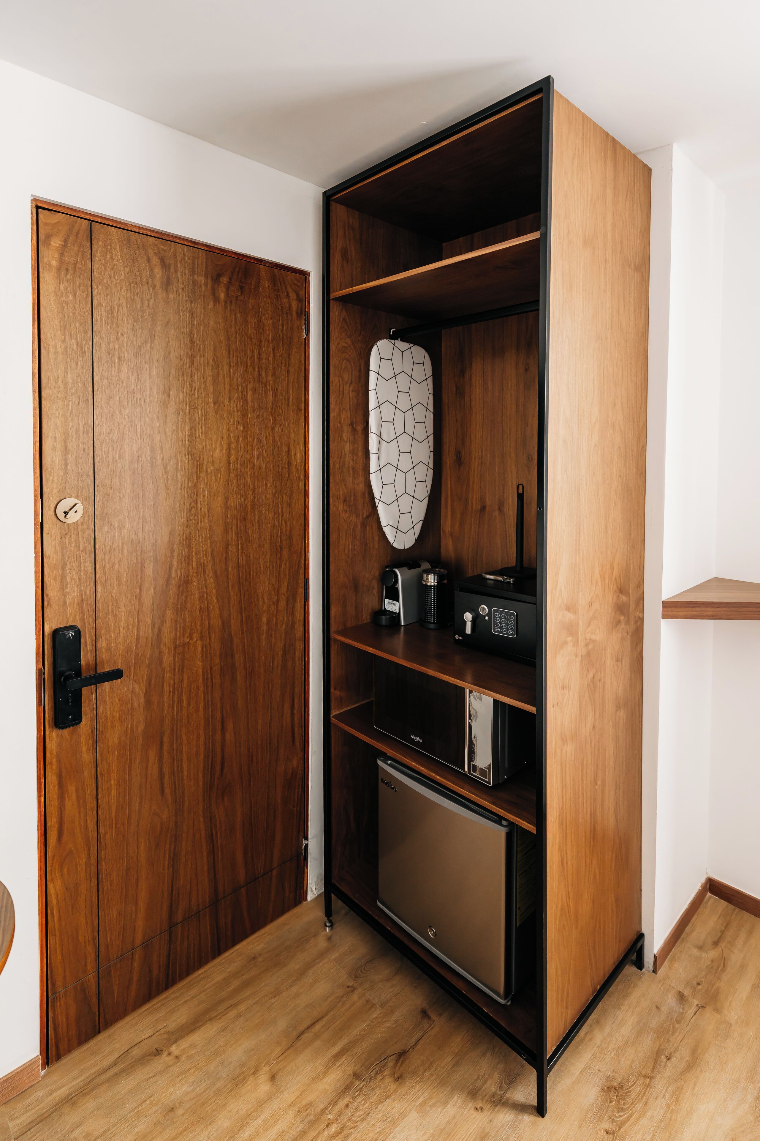 A wooden wardrobe with a black handle, a geometric-patterned mirror, and a black appliance on the shelves.