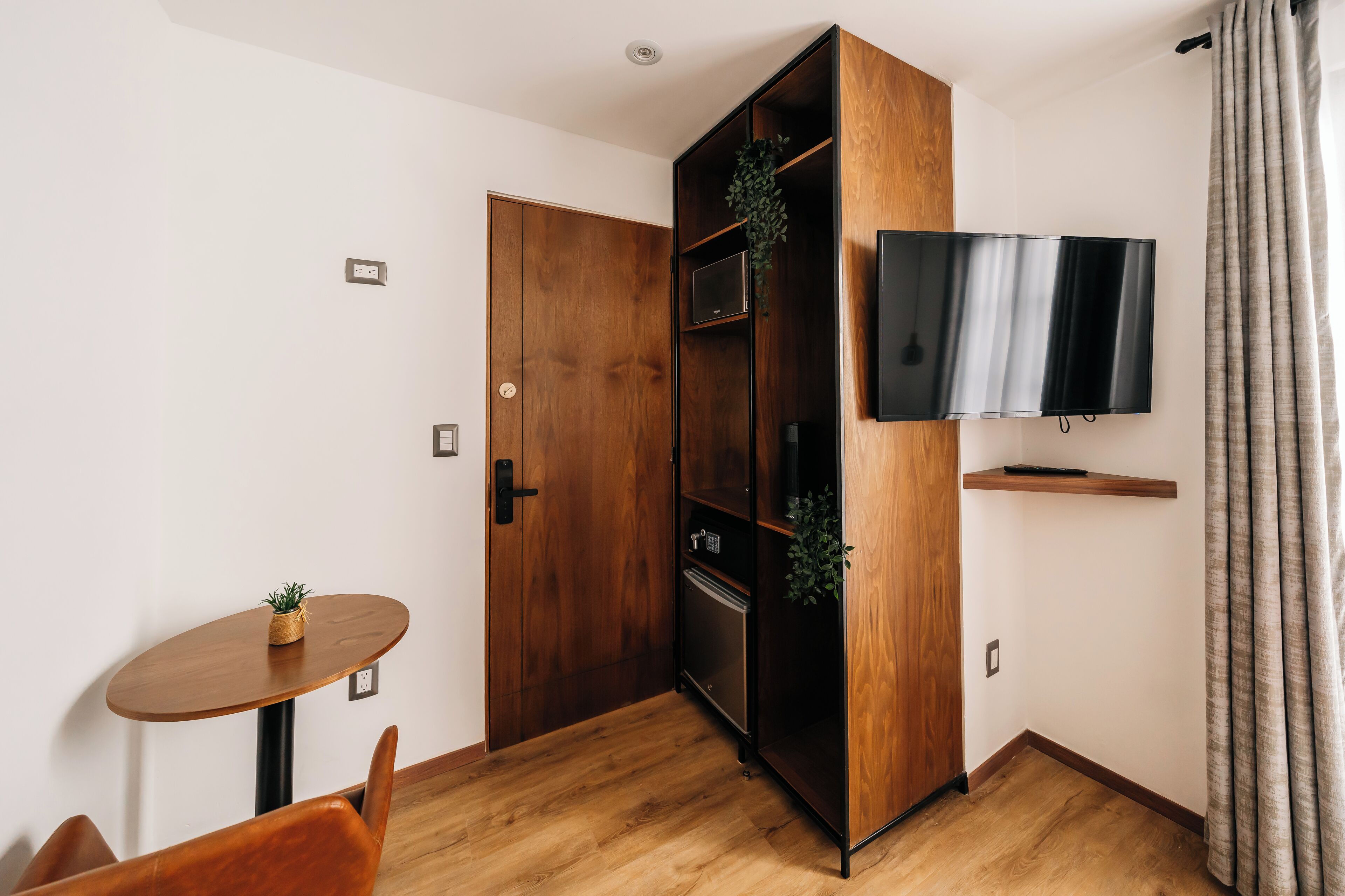 A compact room with a wooden wardrobe, a small table with a plant, a TV mounted on the wall, and a door.