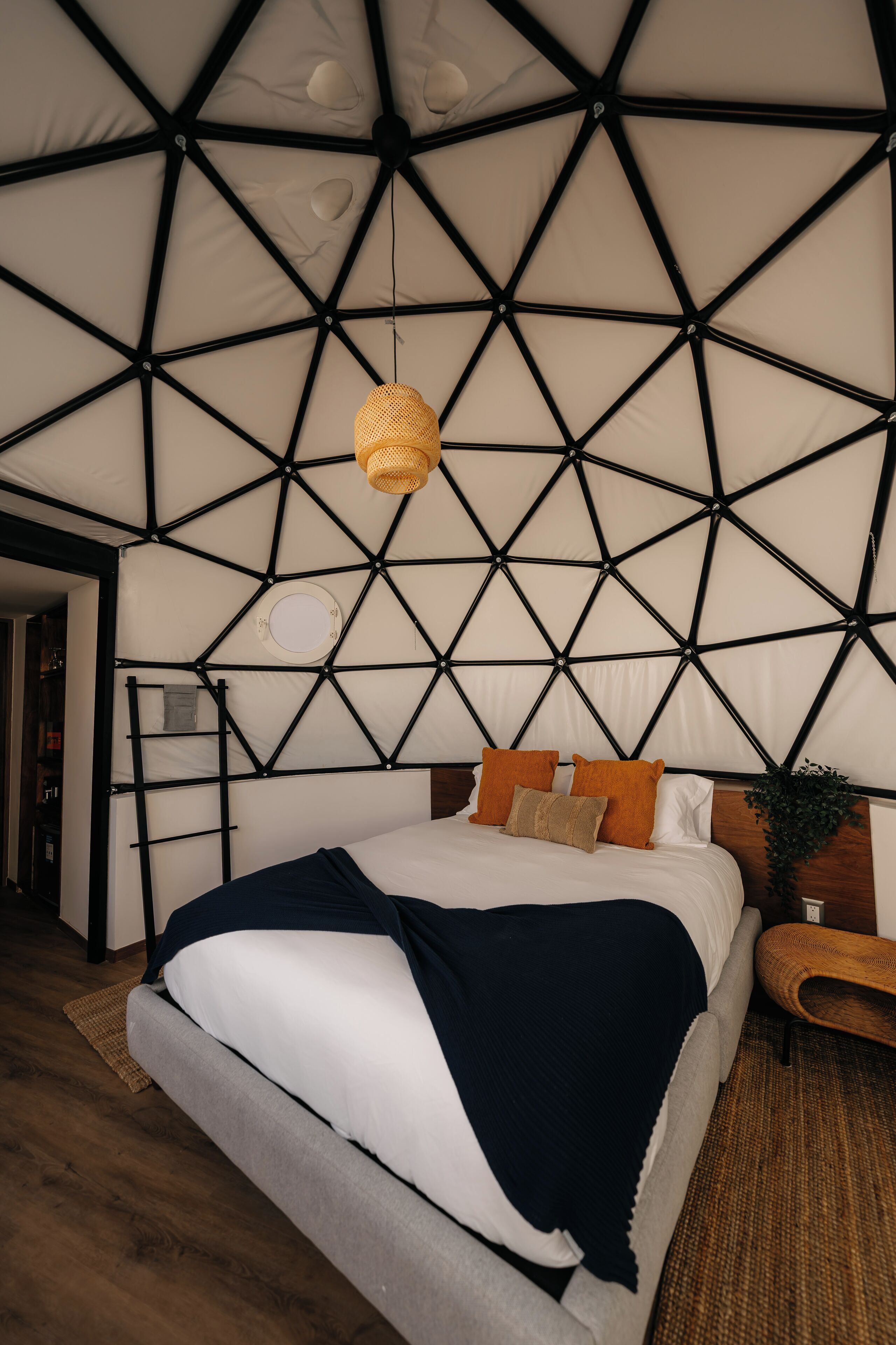 Un dormitorio moderno con techo de patrón geométrico, cama con ropa de cama blanca y una manta oscura, mesita de noche de madera y una lámpara de fibras.