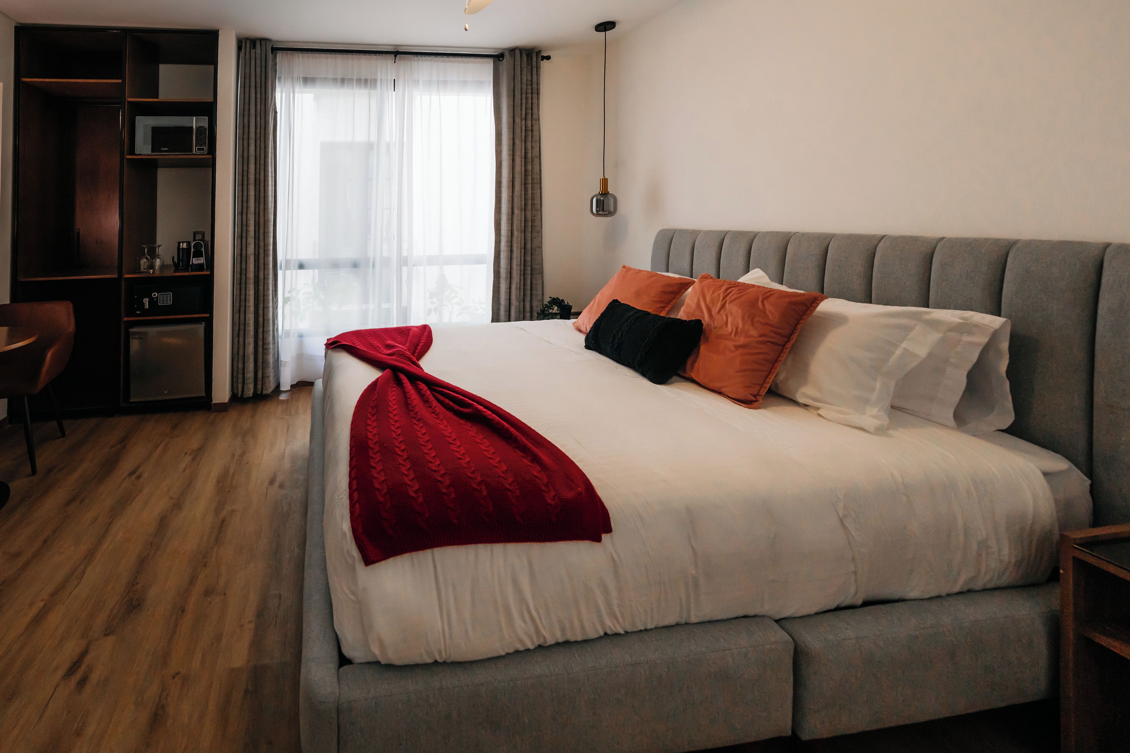 A large bed with a grey headboard, white bedding, and a red throw blanket. There is a wooden wardrobe with a microwave on top.