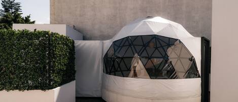 Una estructura moderna con forma de tienda de campaña y fachada de vidrio geométrica, ubicada sobre una terraza de madera junto a una pared blanca y un seto.