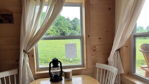 A wooden cabin room with a round table, two white chairs, a lantern, and a window with a view of a green field.