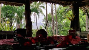 Un cadre tropical avec un toit de chaume, une table recouverte d’une nappe rouge et une vue sur des palmiers et des montagnes.