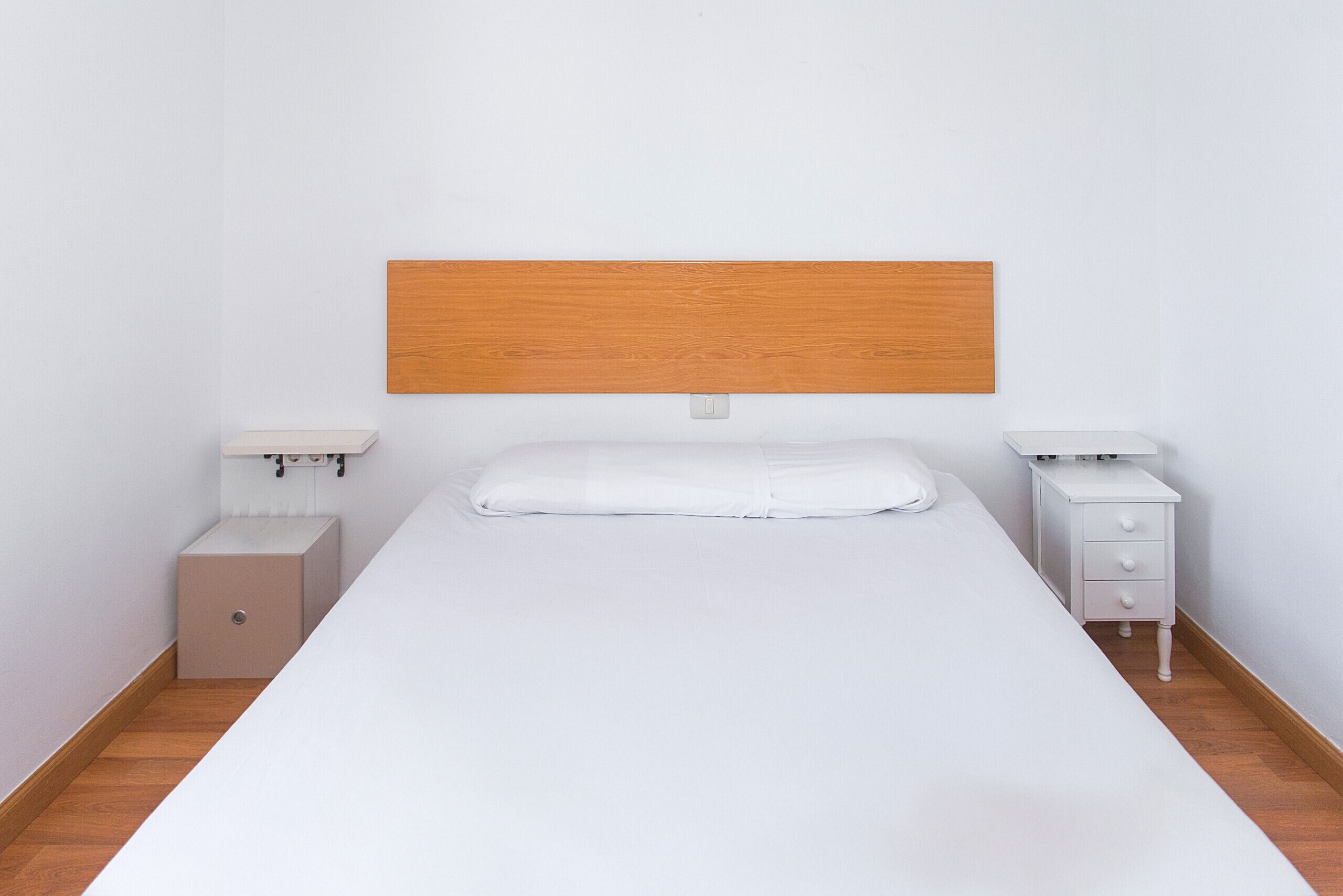 Un dormitorio minimalista con cabecera de madera, una cama individual, dos mesitas de noche y un armario pequeño.