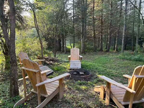 Dos sillas Adirondack mirando hacia un brasero en un área boscosa.