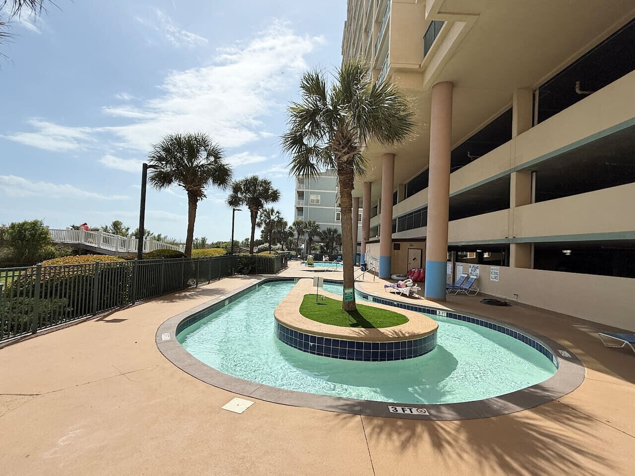 A swimming pool with a palm tree in the center, surrounded by a concrete deck and a multi-story parking garage in the background.