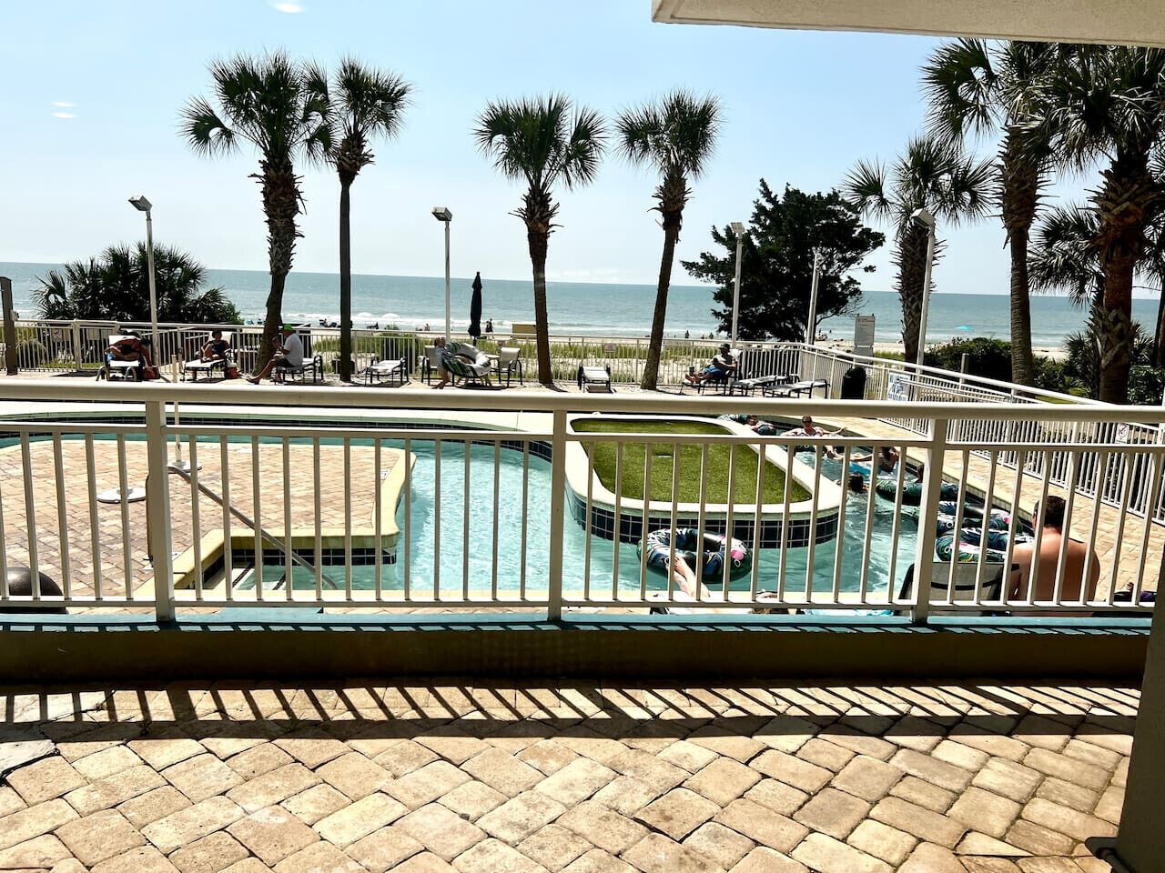 A pool area with a view of the beach, palm trees, and a fenced deck.