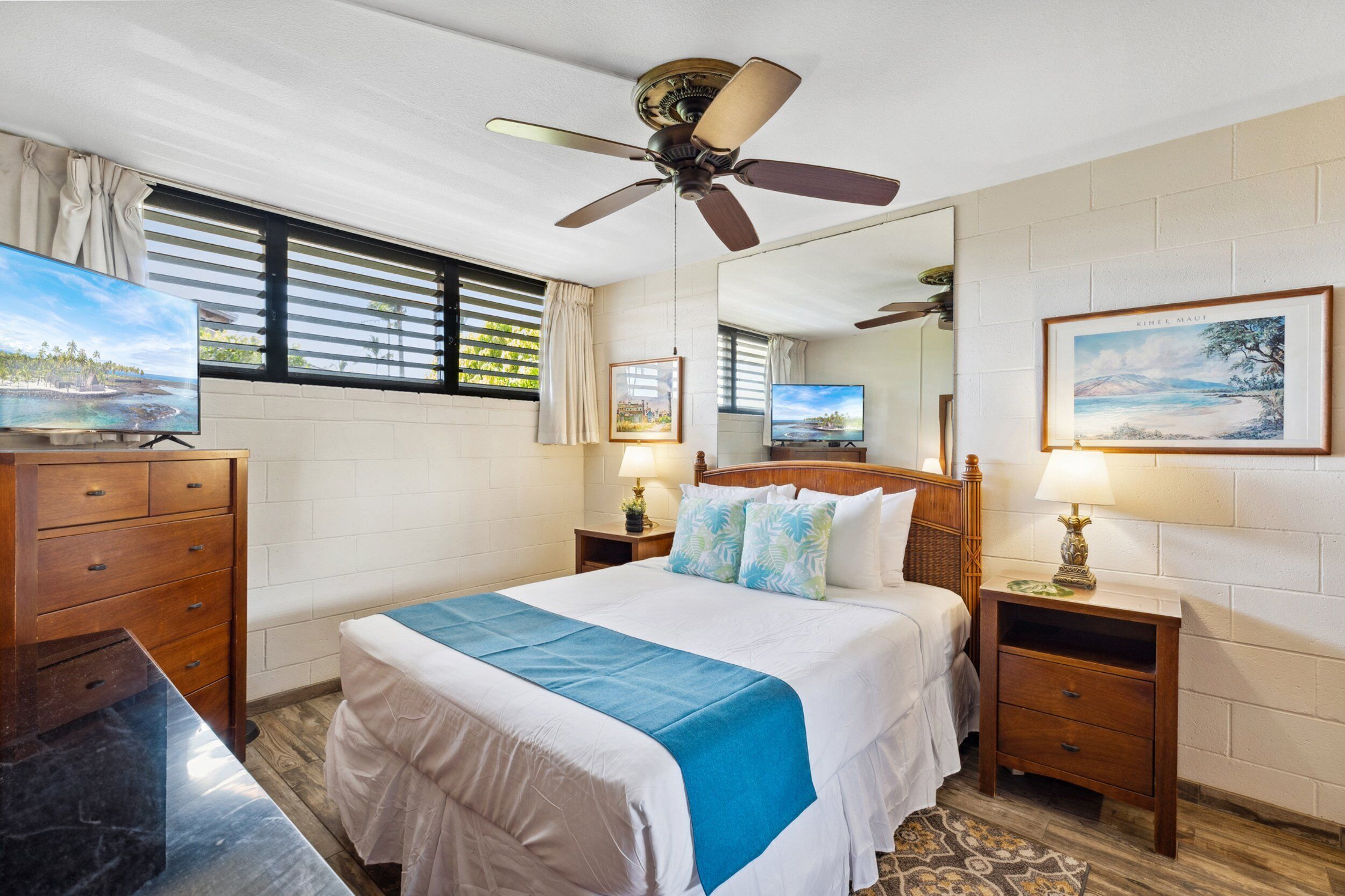 Une chambre d’hôtel avec un lit, un ventilateur de plafond, une télévision, une commode en bois, une table de chevet avec une lampe et un tableau encadré au mur.