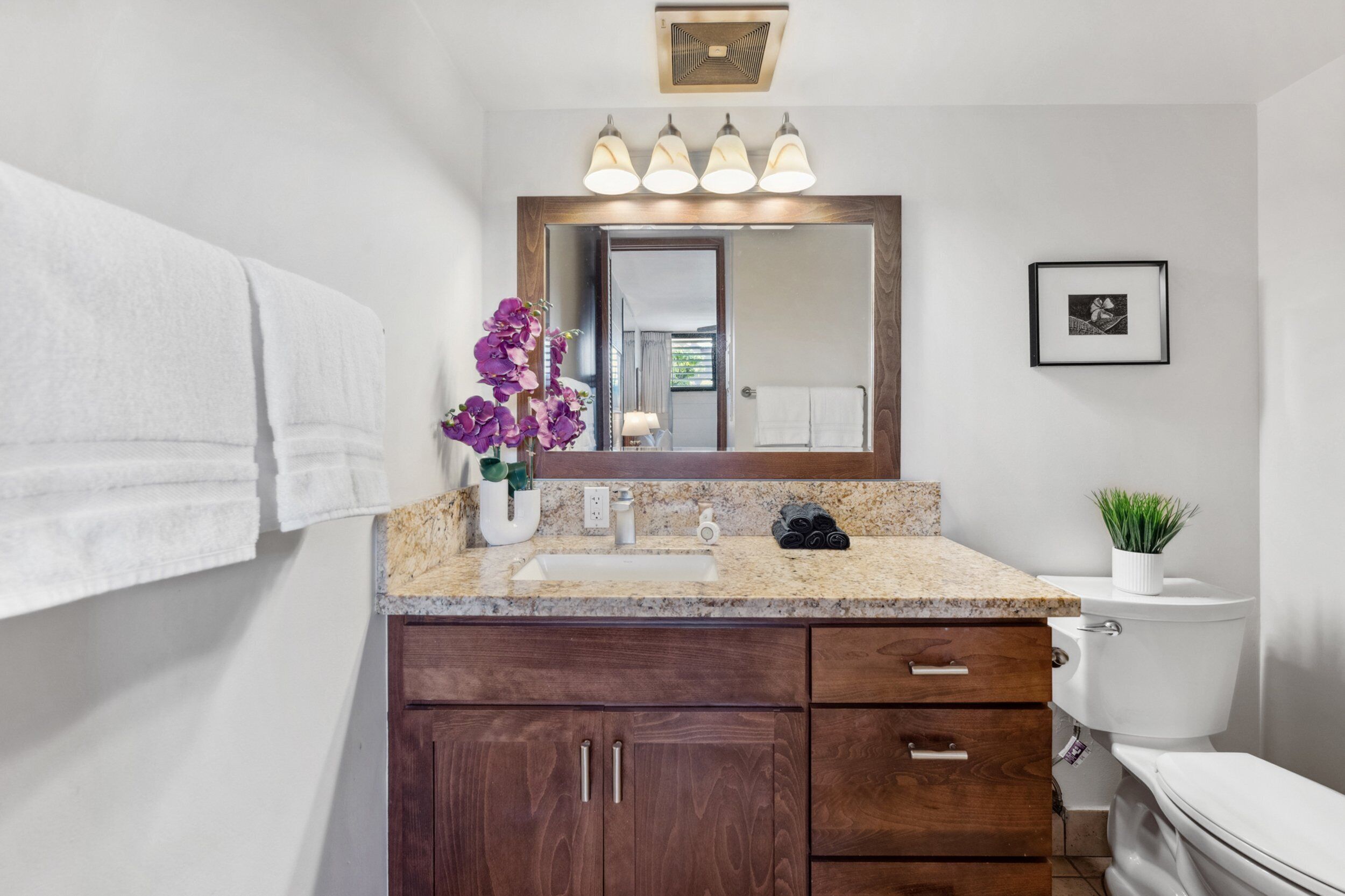 A bathroom with a white sink, dark wooden cabinets, a mirror with three lights, a toilet, and a framed picture on the wall.