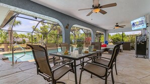 Une terrasse extérieure avec une table en verre, des chaises, un ventilateur de plafond et une piscine en arrière-plan.