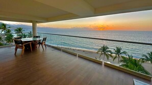 Un balcón con vista al océano al atardecer, piso de madera y una mesa con sillas.
