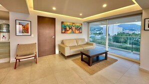 Una sala moderna con un sofá, una mesa de centro y una silla de mimbre. Hay un cuadro en la pared y una puerta que da a otra habitación.
