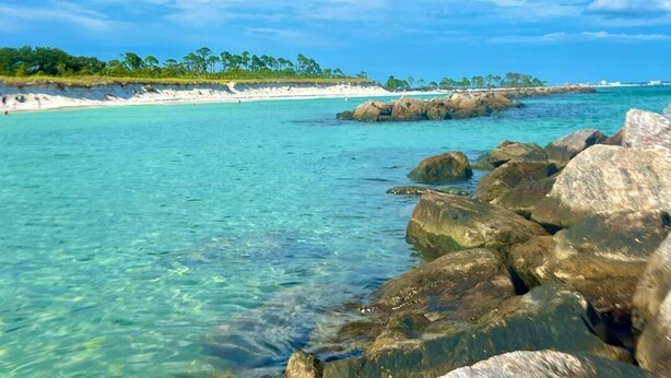 Une scène de plage côtière avec une eau turquoise et limpide, de gros rochers au premier plan et une plage de sable avec des arbres en arrière-plan.