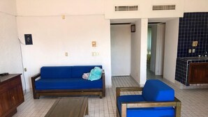 A room with a blue sofa, a wooden chair, and a small table. There are tiled floors and a kitchen area in the background. - Perfect suite for your vacation (Ixtapa Zihuatanejo)
