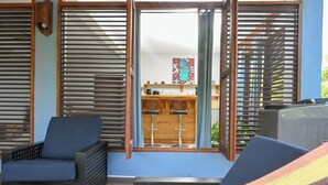 Une pièce comprenant un canapé, un fauteuil, un coin bar avec des tabourets et une fenêtre avec des stores, offrant une vue sur l’extérieur.