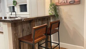 Une cuisine dotée d’un îlot central en bois, de deux tabourets de bar, de luminaires suspendus, d’une fenêtre avec stores et d’une étagère murale où sont rangés les ustensiles de cuisine.
