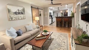 Un salon avec un canapé, une table basse et un tapis. Au fond, on aperçoit un coin cuisine avec des placards et un plan de travail.