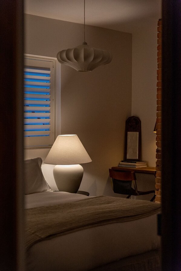 Un dormitorio con cama, lámpara de noche, ventana con persianas e iluminación colgante.