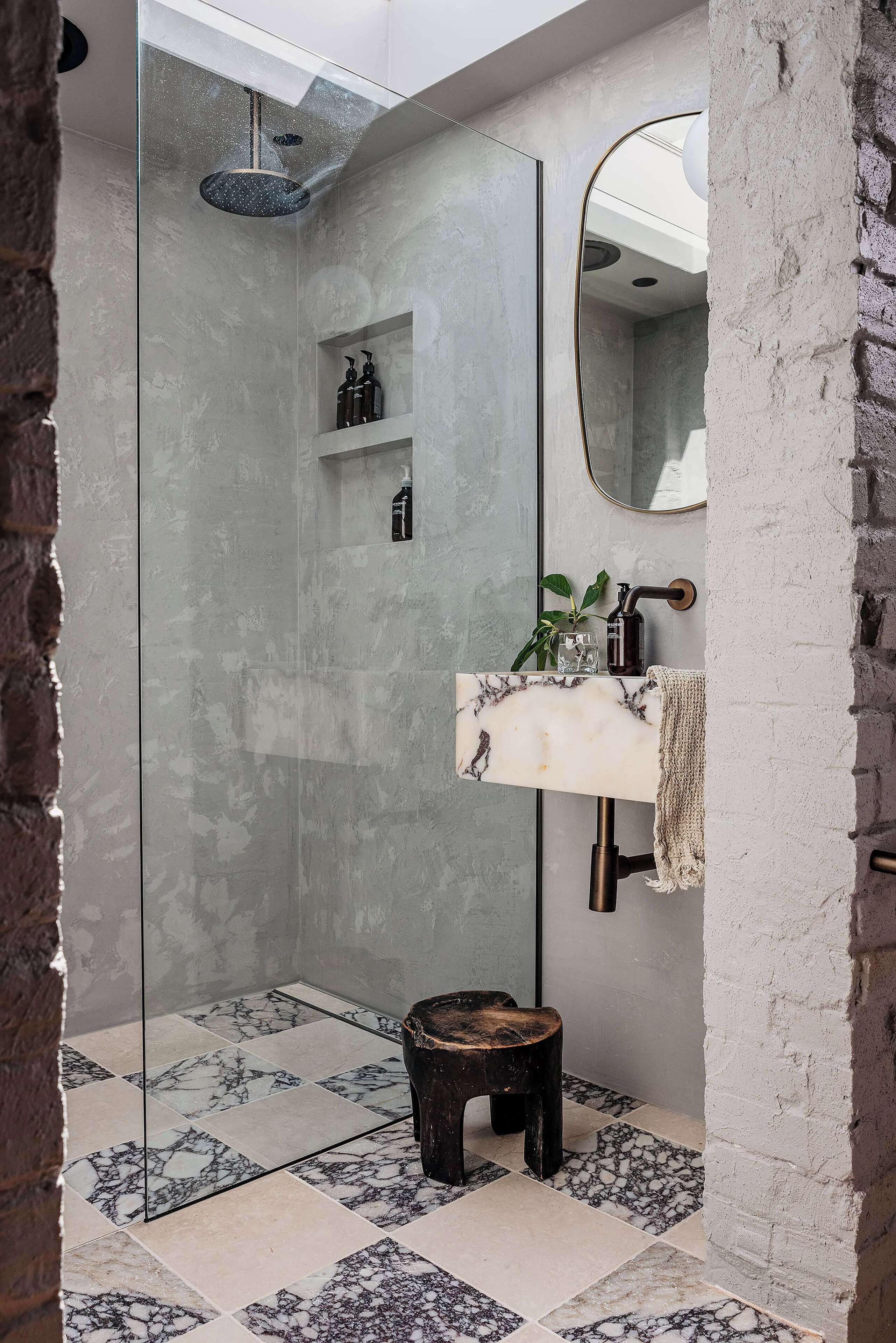 A bathroom with a glass-enclosed shower, marble-patterned floor, a wooden stool, a sink with a towel, and a mirror.
