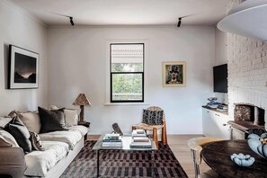 A living room with a sofa, a coffee table with books, a chair, a fireplace, and a large window. - The Top Floor at Collected Places Hobart (North Hobart)