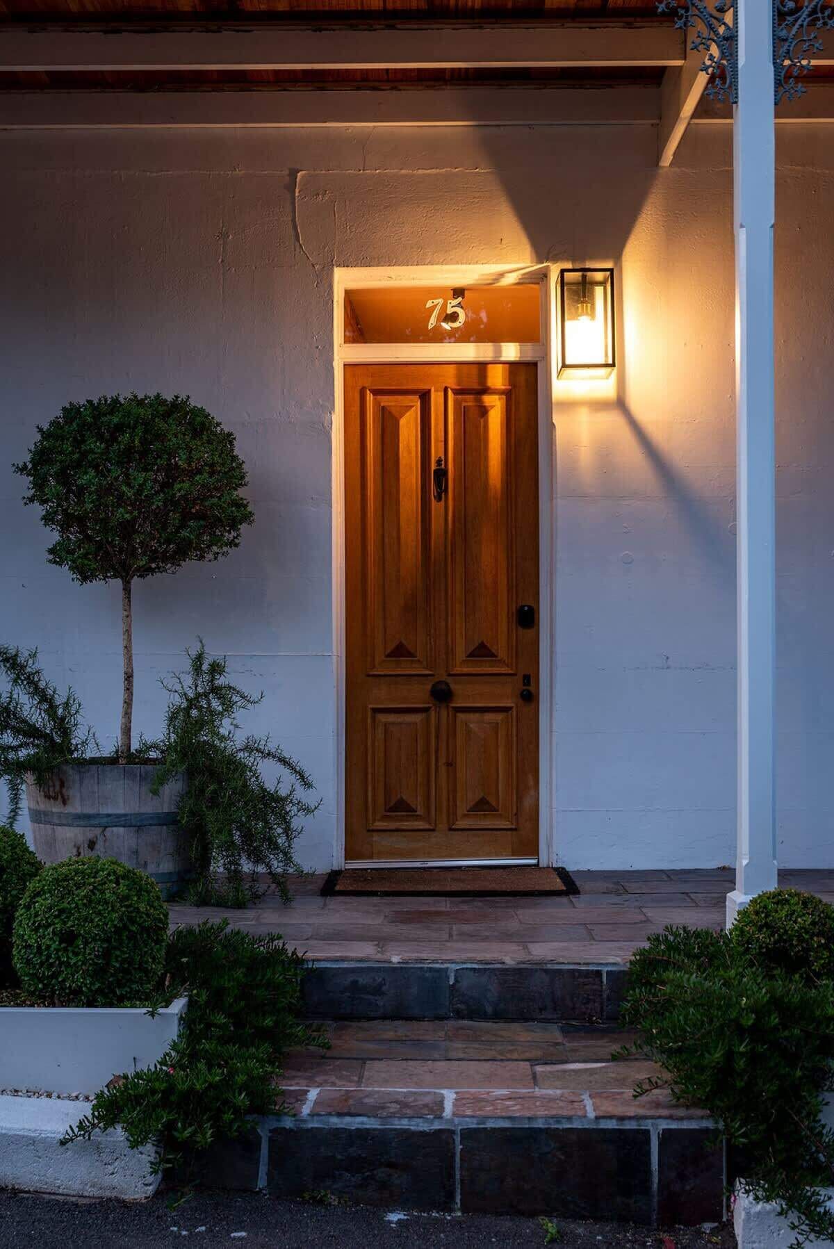 Una puerta de madera con el número 75 arriba, un pequeño porche con escalones y un árbol en maceta a la izquierda.