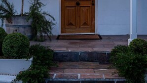 Eine Holztür mit der Nummer 75 darüber, eine kleine Veranda mit Stufen und ein Blumentopf mit einem Baum links.