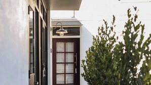A narrow pathway leading to a door with a glass panel, flanked by potted plants and a hanging light fixture.
