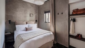 A bedroom with a bed, a hanging light fixture, a window with curtains, a coat rack with a jacket, and a pair of boots.