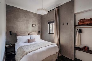 A bedroom with a bed, a hanging light fixture, a window with curtains, a coat rack with a jacket, and a pair of boots.