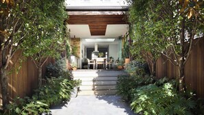 A stone pathway leading to a wooden deck with a dining area, surrounded by lush greenery and a wooden fence.
