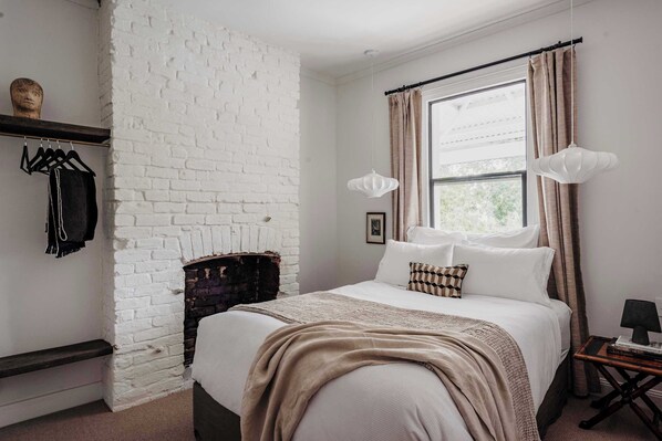 Quarto com parede de tijolos brancos, cama com roupa de cama bege, janela com cortinas e luminária pendente.