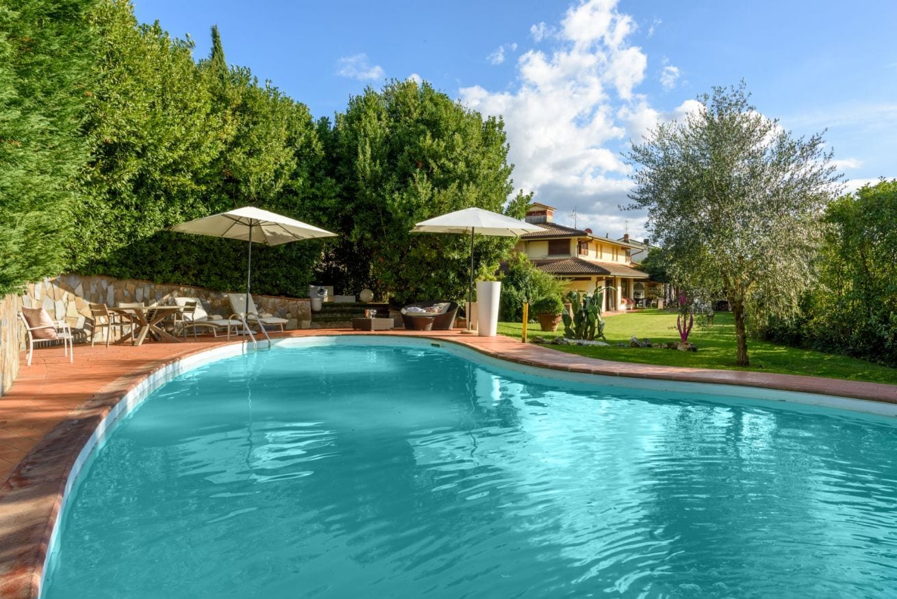Une piscine entourée de verdure et d’une maison, avec des chaises longues et des parasols.