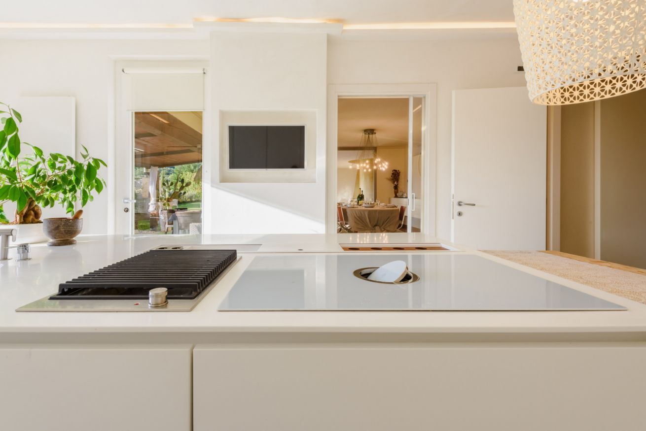 A modern kitchen with a central island, a flat-screen TV mounted above it, and a dining area visible through an open door.