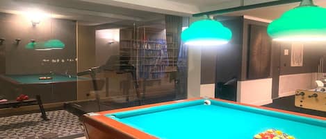 Una mesa de billar con superficie de fieltro verde, una partición de vidrio y una estantería al fondo.