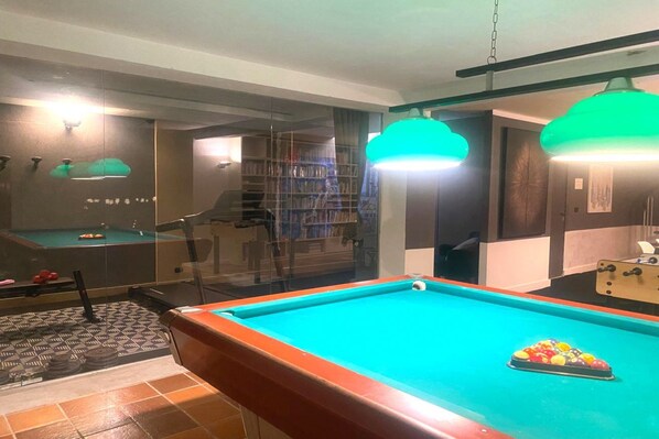 Une table de billard recouverte d’un drap vert, une cloison en verre et une bibliothèque en arrière-plan.